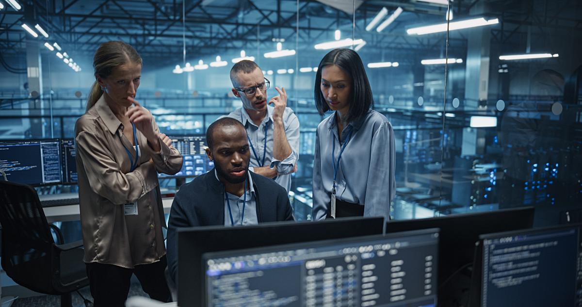 Diverse Team of IT Professionals Gathered Behind a Desk, Looking at Data on Computer Screens, Problem Solving a Data Security Project, Brainstorming Future Upgrades for Cloud Computing