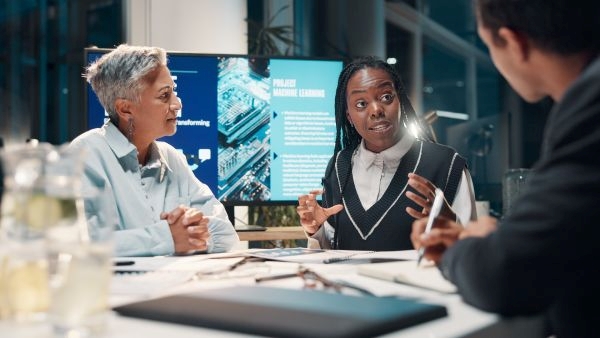 A diverse team of professionals discussing IT consulting strategies in a modern office with technology screens in the background.
