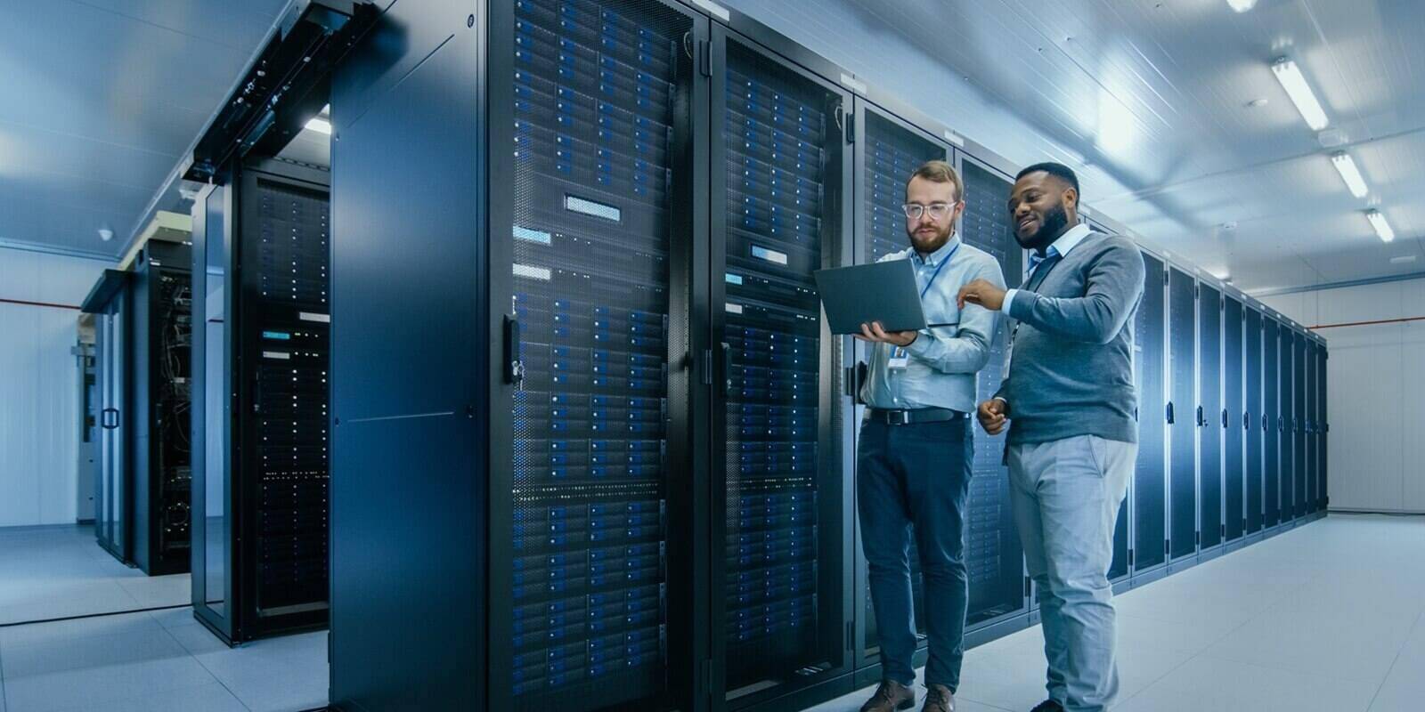 Bearded IT Technician in Glasses with a Laptop Computer and Black Male Engineer Colleague are Talking in Data Center while Working Next to Server Racks
