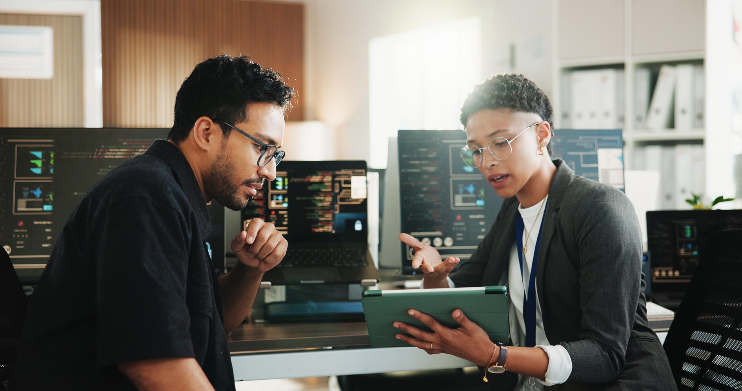 IT professionals collaborating over a tablet with code and system monitors in the background