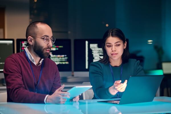 young woman as female IT security expert using laptop with male colleague in blue neon light